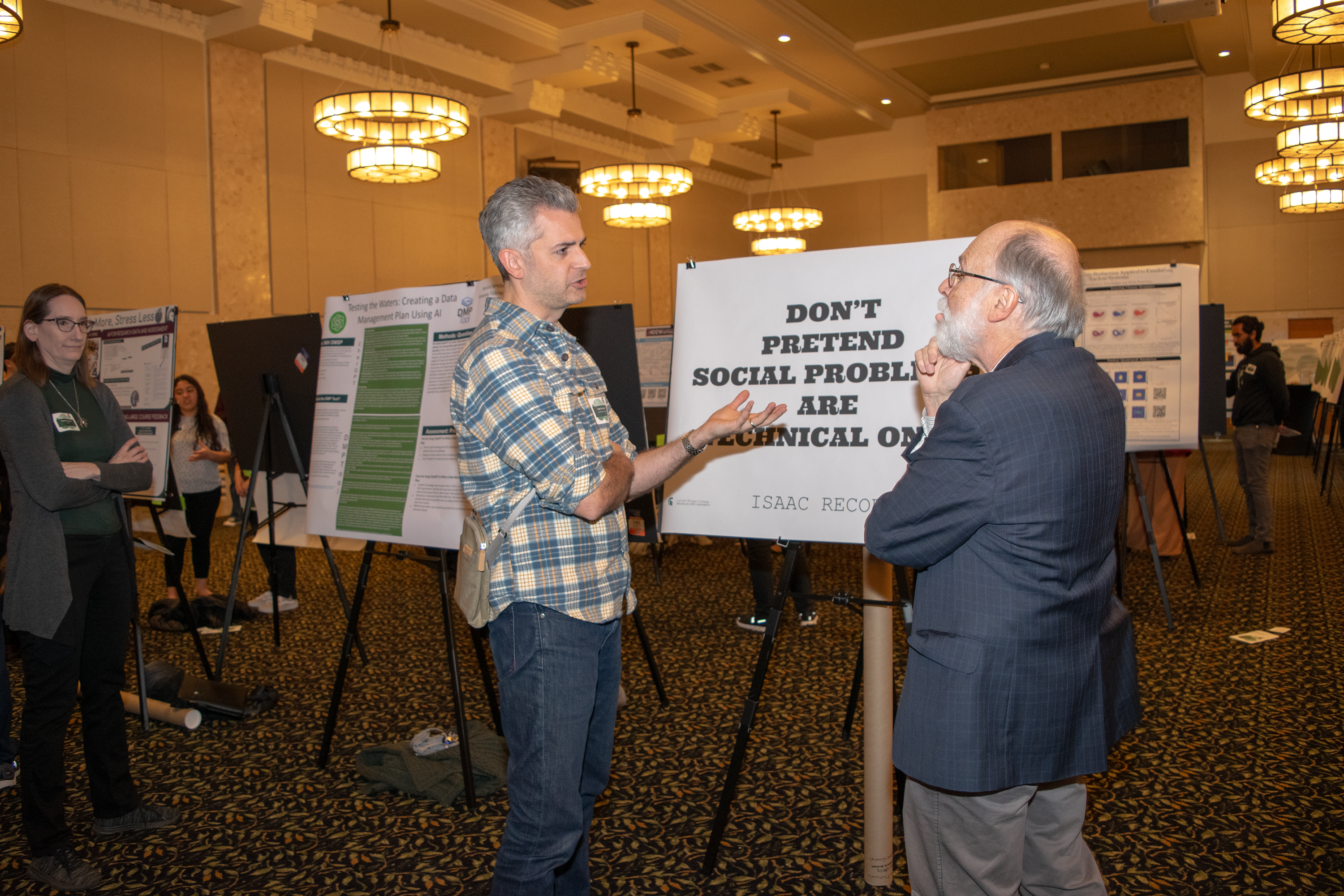 A man in a plaid shirt talks to a man with a beard and glasses in a suit in front of a research poster